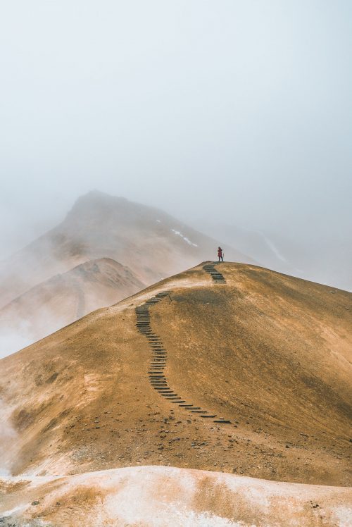 Foto de un hombre en la cima de un cerro con un sendero. Detrás se ve otro cerro más alto, entre la neblina.