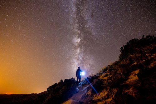 Foto de un joven sosteniendo un farol en un camino ; a su izquierda el amanecer y a su derecha la noche estrellada.