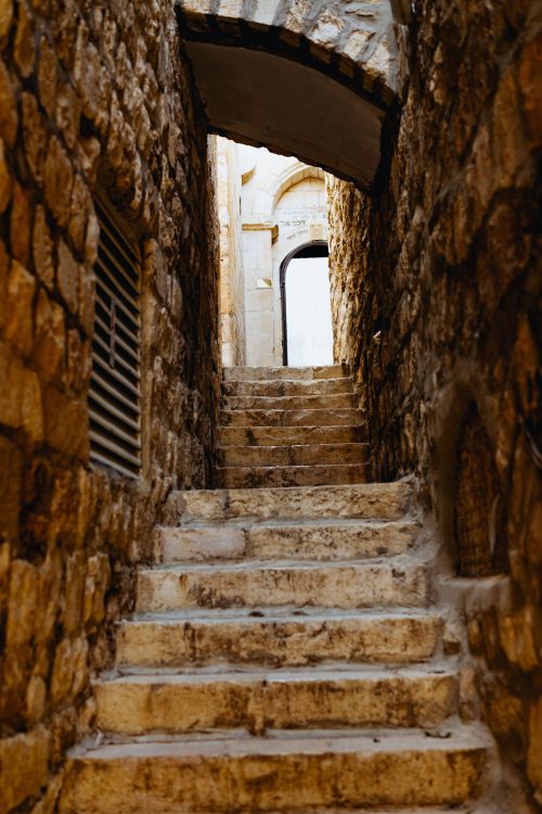 Una escalinata en un pasillo angosto que conduce a una puerta abierta donde se observa el cielo
