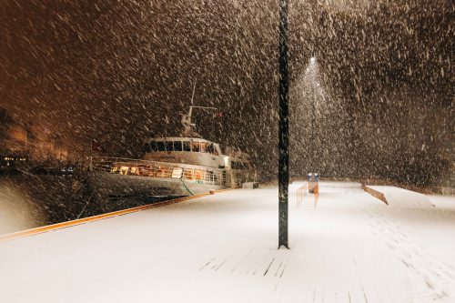 Foto de un barco anclado a un muelle iluminado en la noche, mientras cae una tormenta de nieve